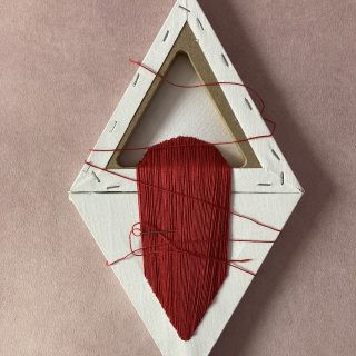 Zdjęcie pionowe. Praca artystyczna na jasnoróżowym tle. Składa się z dwóch białych trójkątnych płócien pod obrazy złączonych ze sobą jednym z boków tworząc pionowy romb. Trójkątne płótno u góry jest ukazane tyłem, dolne przodem. Z górnego wychodzi szeroki haftowany pas czerwoną nicią i wchodzi ponownie w dolny trójkąt, przypominając tym samym krwisty wodospad. Pozostała czerwona nitka luźno oplata cały romb, a igła jest wetknięta w wyhaftowany pas.